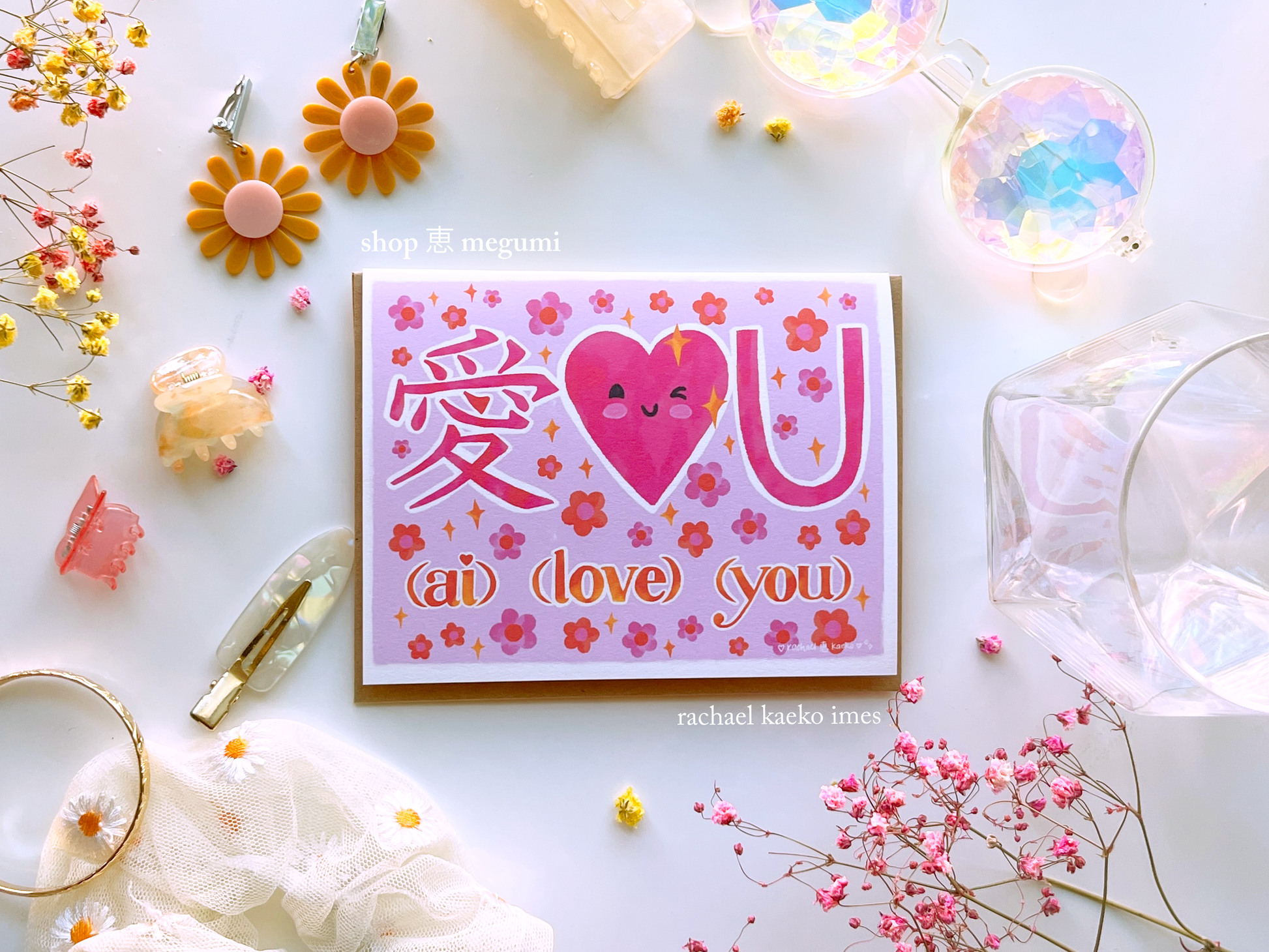Hand-drawn greeting card featuring pink illustrated text that reads "Ai Love You" with the Japanese kanji character for love, a smiling pink heart, and pink daisy flowers and yellow sparkles, on a white surface surrounded by colorful objects.