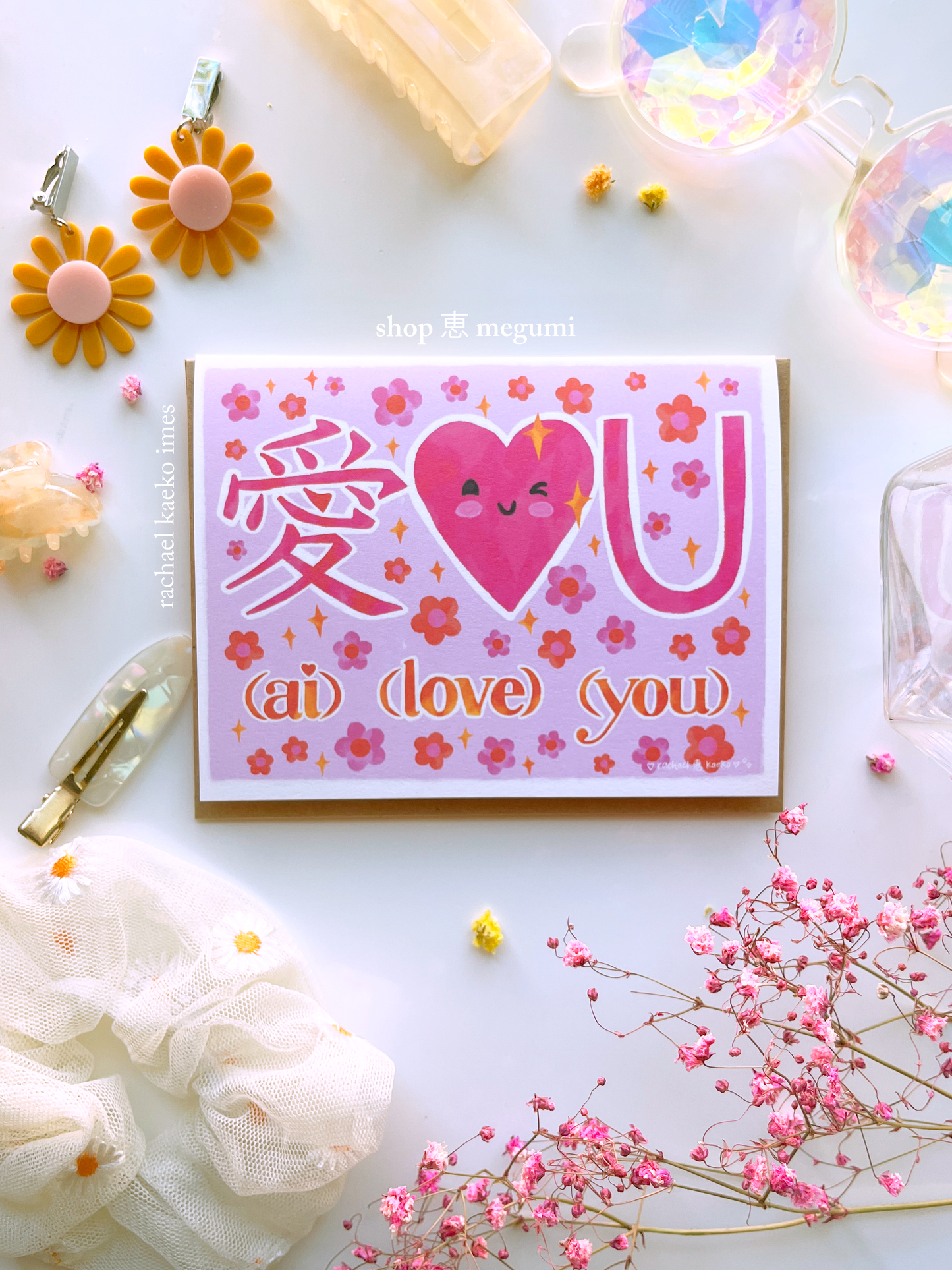 Hand-drawn greeting card featuring pink illustrated text that reads "Ai Love You" with the Japanese kanji character for love, a smiling pink heart, and pink daisy flowers and yellow sparkles, on a white surface surrounded by colorful objects.