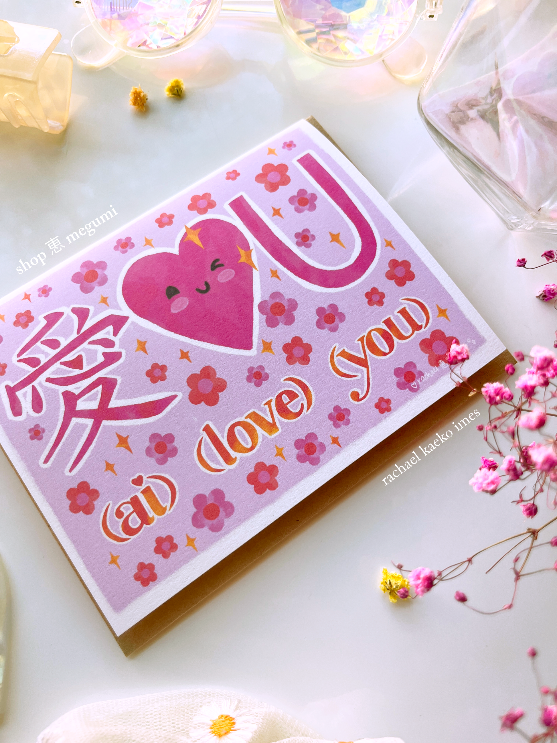 Hand-drawn greeting card featuring pink illustrated text that reads "Ai Love You" with the Japanese kanji character for love, a smiling pink heart, and pink daisy flowers and yellow sparkles, on a white surface surrounded by colorful objects.
