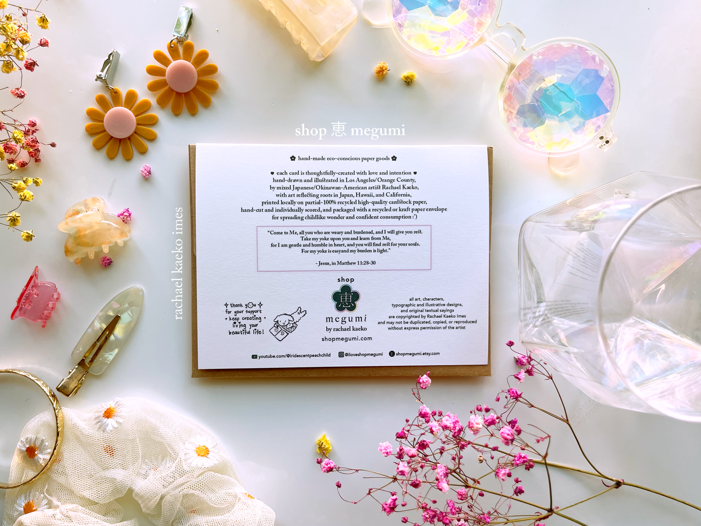 Back view of greeting card with shop megumi branding information and logo, on a white surface with colorful items.