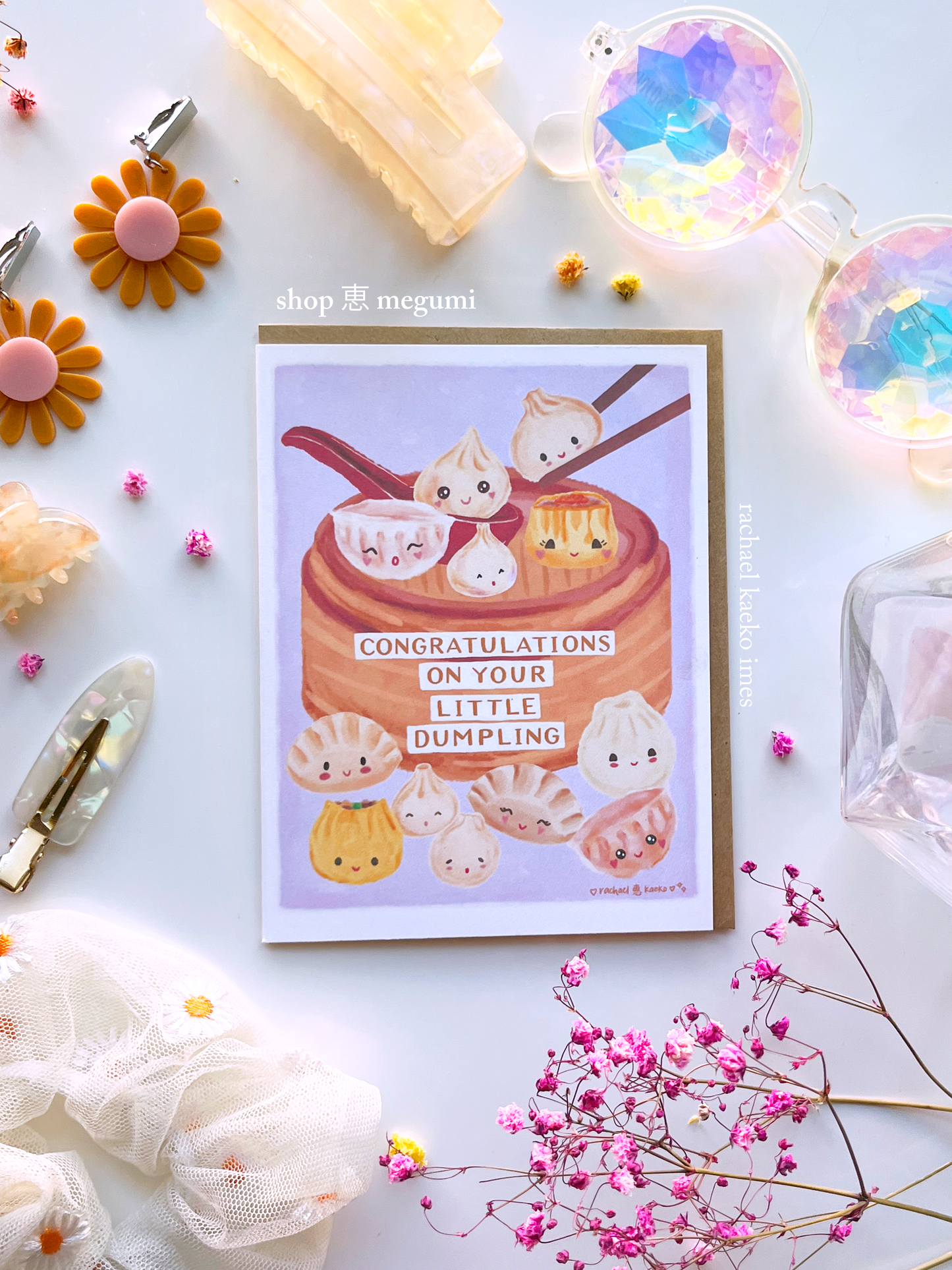 Greeting card with illustration of dumplings in a bamboo basket with "Congratulations on Your Little Dumplings" text, on a light background, surrounded by decorative items.