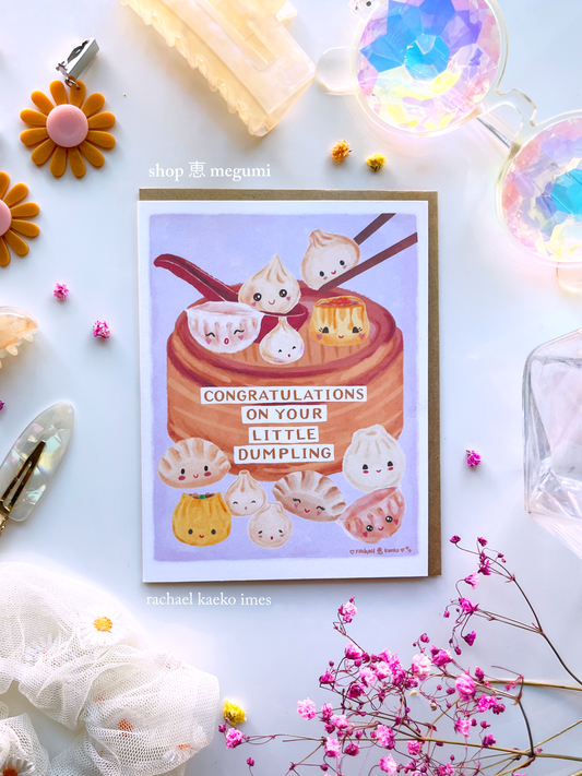 Greeting card with illustration of dumplings in a bamboo basket with "Congratulations on Your Little Dumplings" text, on a light background, surrounded by decorative items.