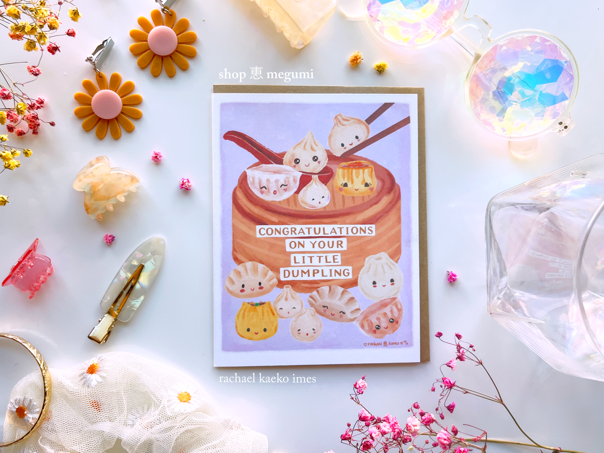 Greeting card with illustration of dumplings in a bamboo basket with "Congratulations on Your Little Dumplings" text, on a light background, surrounded by decorative items.