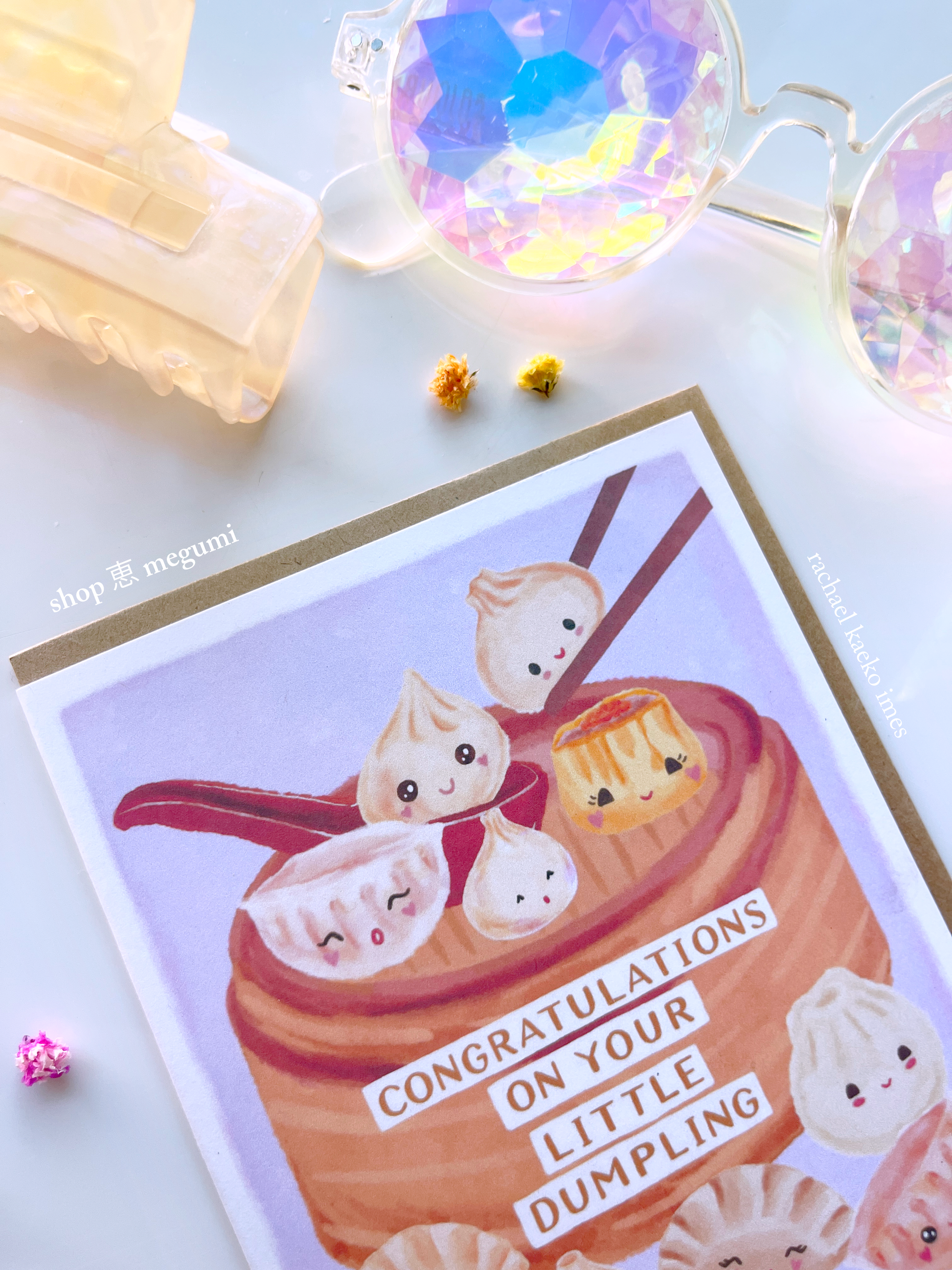 Close-up of greeting card with illustration of dumplings in a bamboo basket with "Congratulations on Your Little Dumplings" text, on a light background, surrounded by decorative items.