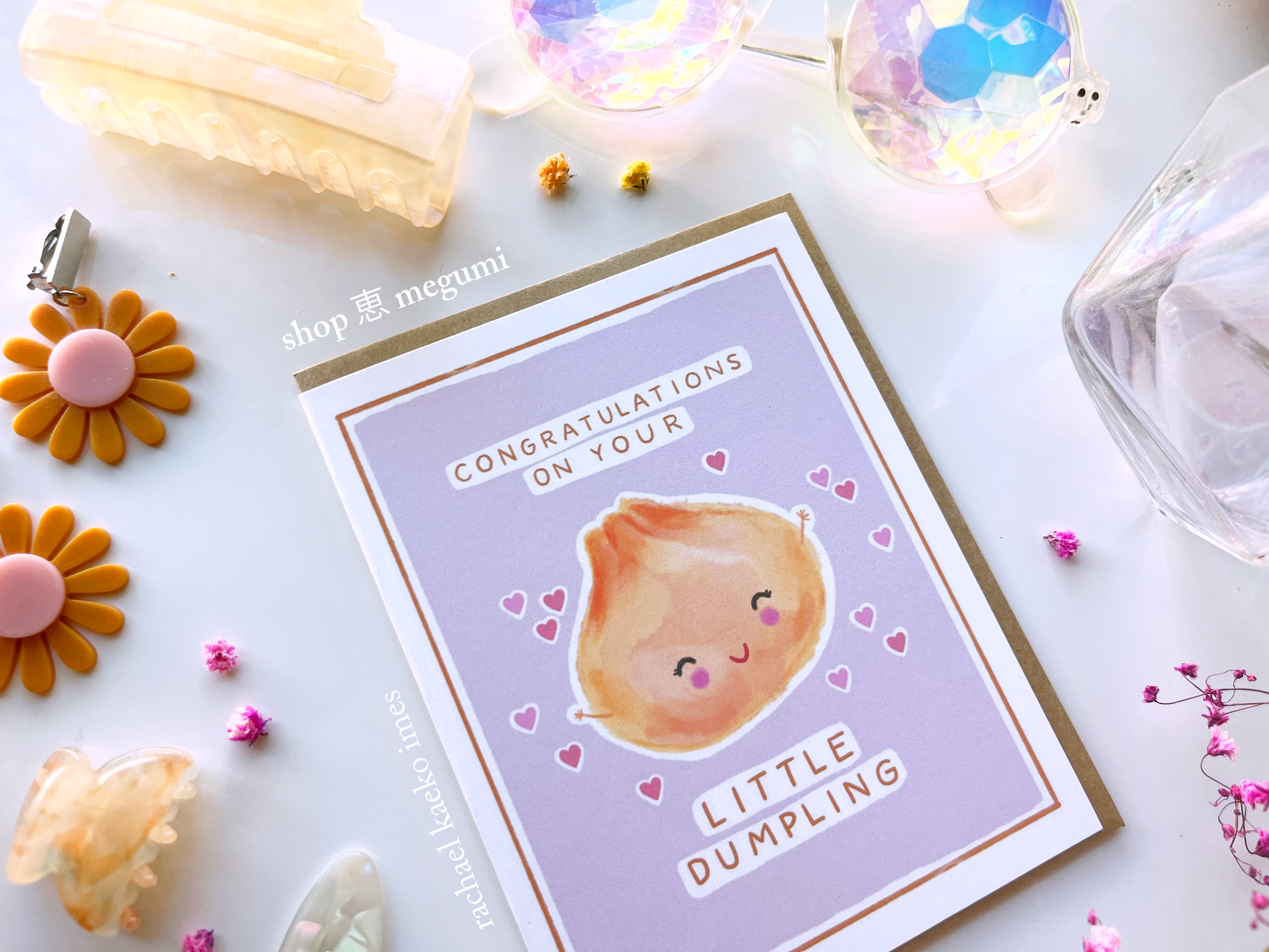 Close-up image of greeting card with a cute illustration of a dumpling and text that reads "Congratulations on Your Little Dumpling" on a light background with decorative items.