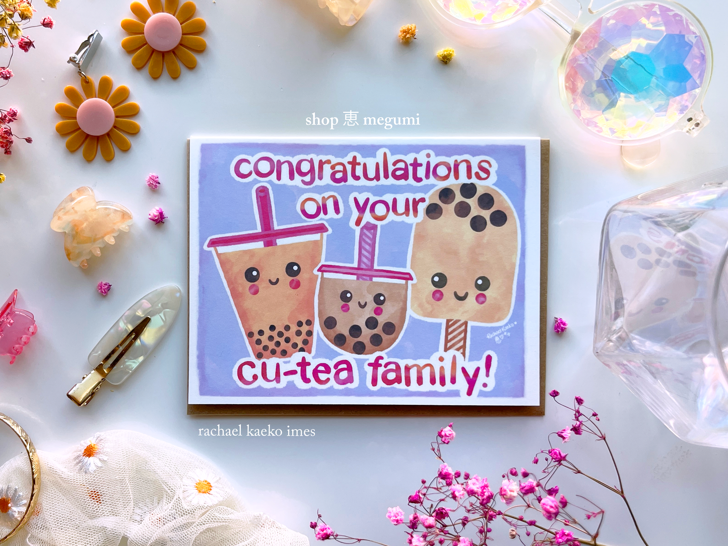 Milk tea-inspired baby shower greeting card featuring illustrations of smiling milk tea bobas with hand-written text that reads "Congratulations on Your Cu-tea Family", on a white surface surrounded by colorful items.