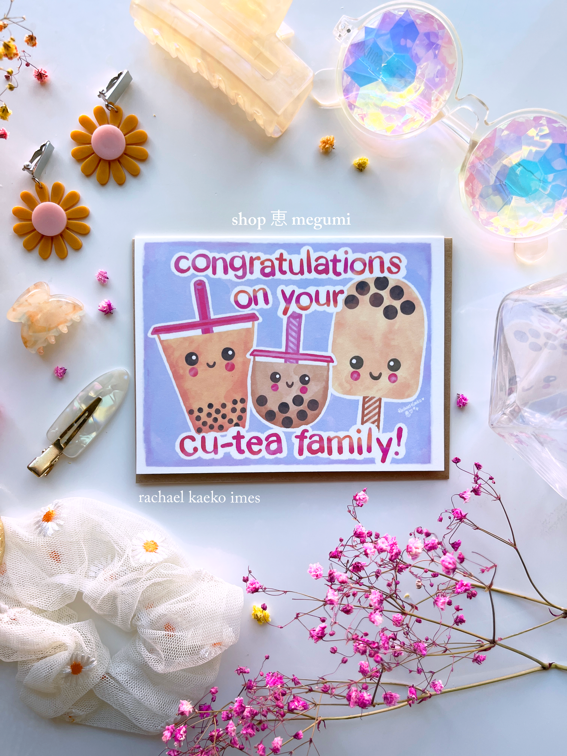 Milk tea-inspired baby shower greeting card featuring illustrations of smiling milk tea bobas with hand-written text that reads "Congratulations on Your Cu-tea Family", on a white surface surrounded by colorful items.
