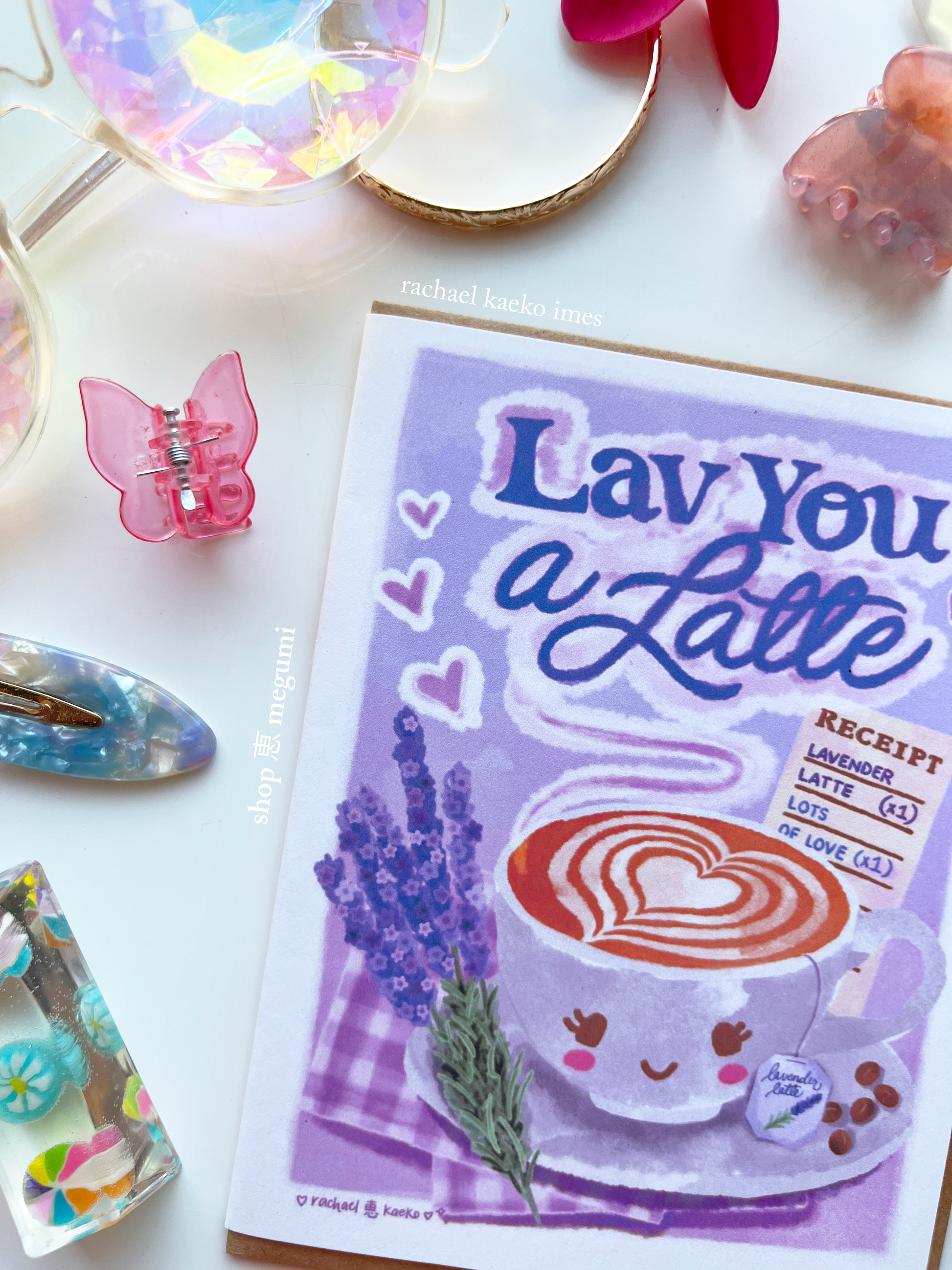 Close-up of a cute coffee shop greeting card with a lavender latte 'Lav You a Latte' design on a white surface with decorative items.