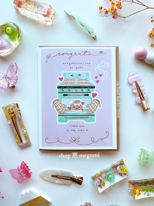 Cute greeting card with a vintage oven mama and cinnamon bun baby illustration surrounded by decorative items on a white background.