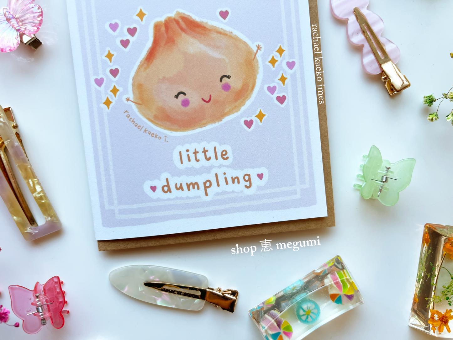 Baby shower greeting card with a cute illustration of a dumpling and text 'little dumpling' surrounded by decorative items on a white surface.