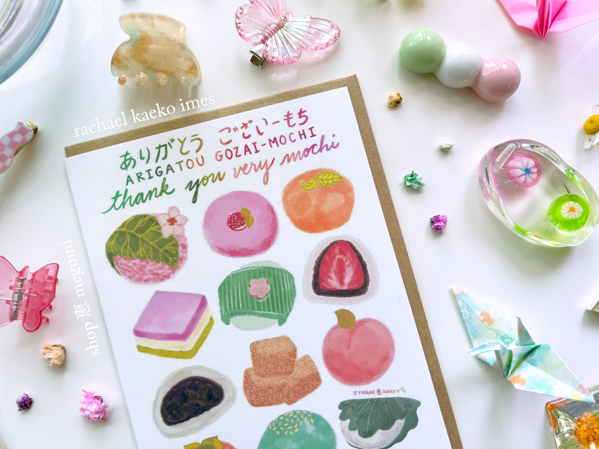 Close-up image of greeting card with illustrations of mochi and text that reads 'Arigatou Gozaimochi' and 'Thank You Very Mochi' in English and Japanese on a white background with decorative items.