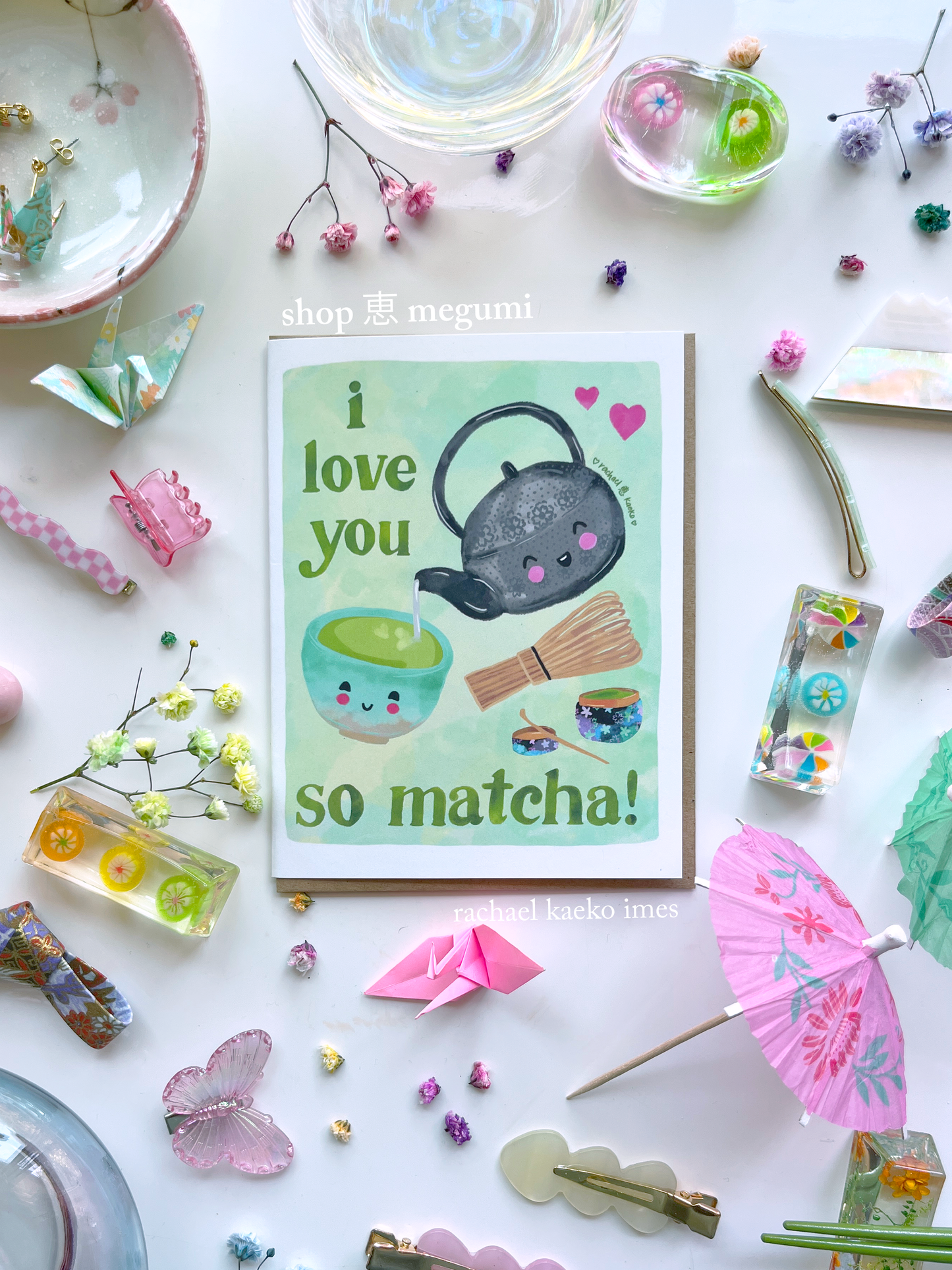 Japanese matcha-inspired greeting card with a smiling teapot and matcha elements, and text that reads "I Love You so Matcha!", on a white surface with decorative items.