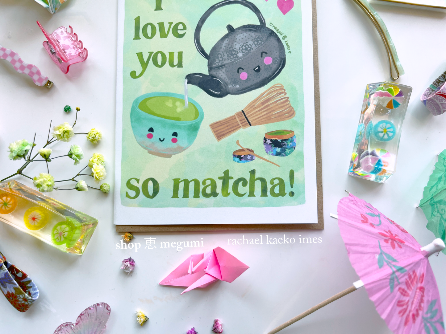 Close-up of Japanese-inspired greeting card with a smiling matcha teacup and matcha elements, on a white surface with decorative items.
