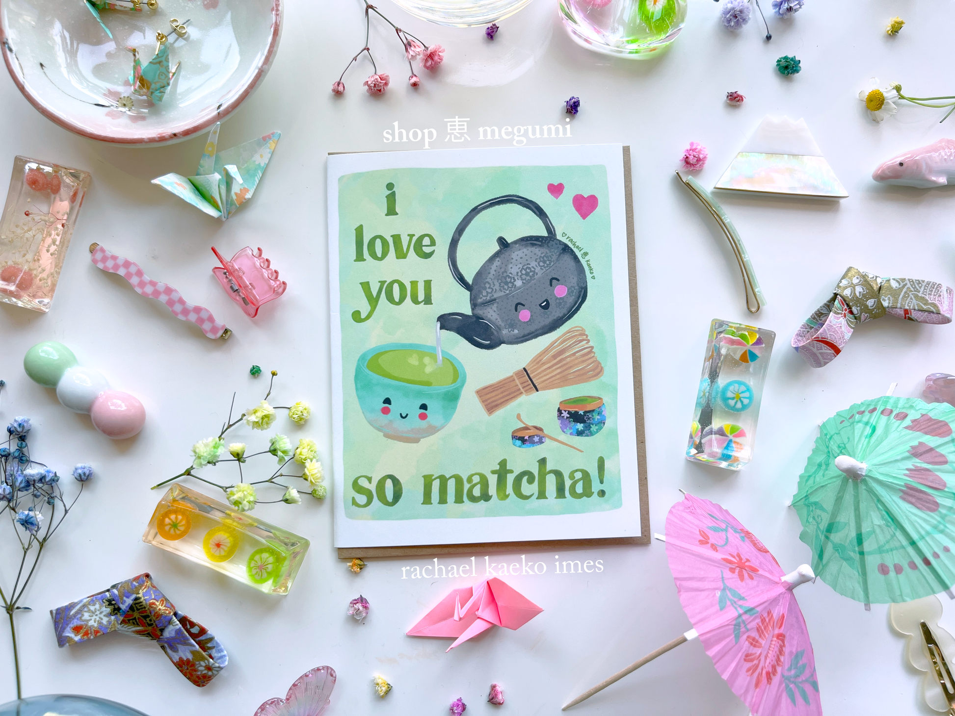 Japanese matcha-inspired greeting card with a smiling teapot and matcha elements, and text that reads "I Love You so Matcha!", on a white surface with decorative items.