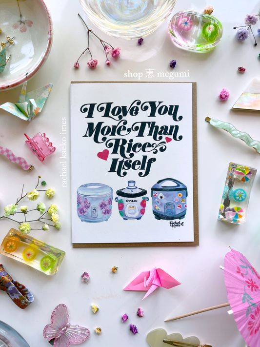 Greeting card with 'I Love You More Than Rice' illustrated text design and colorful illustrations of rice cookers, surrounded by decorative items on a light surface.