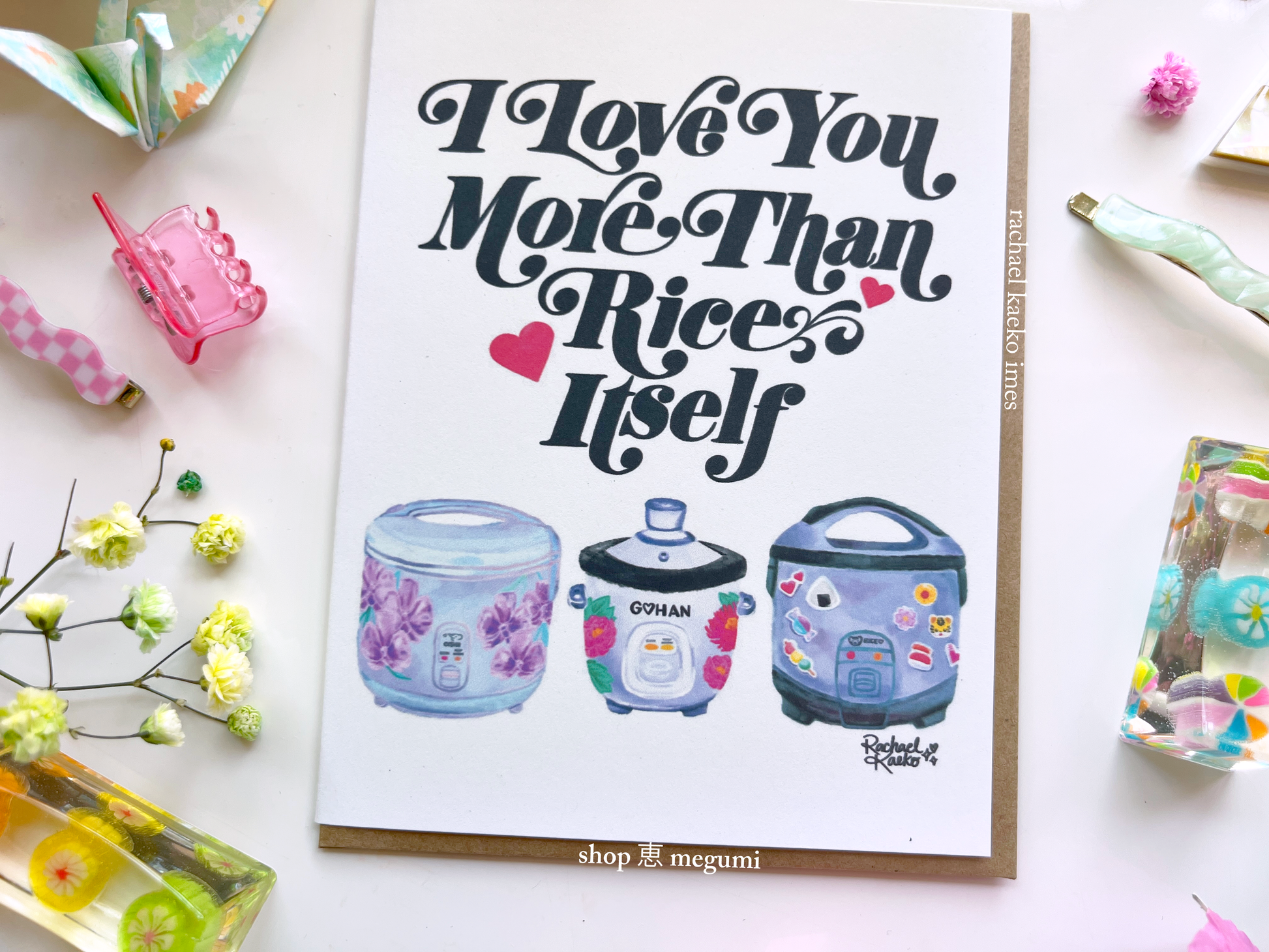 Close-up image of greeting card with 'I Love You More Than Rice' illustrated text design and colorful illustrations of rice cookers, surrounded by decorative items on a light surface.