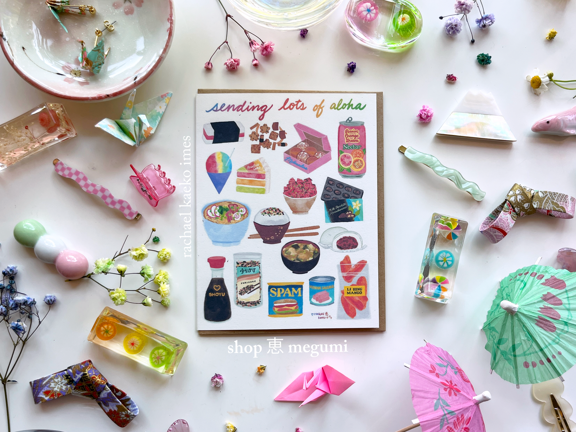 Hawaii-inspired greeting card that reads "Sending Lots of Aloha" in cursive text, with colorful illustrations of Hawaii-style foods, on a white surface with colorful decorative objects.