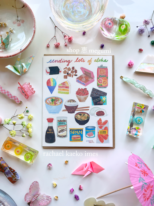 Hawaii-inspired greeting card that reads "Sending Lots of Aloha" in cursive text, with colorful illustrations of Hawaii-style foods, on a white surface with colorful decorative objects.