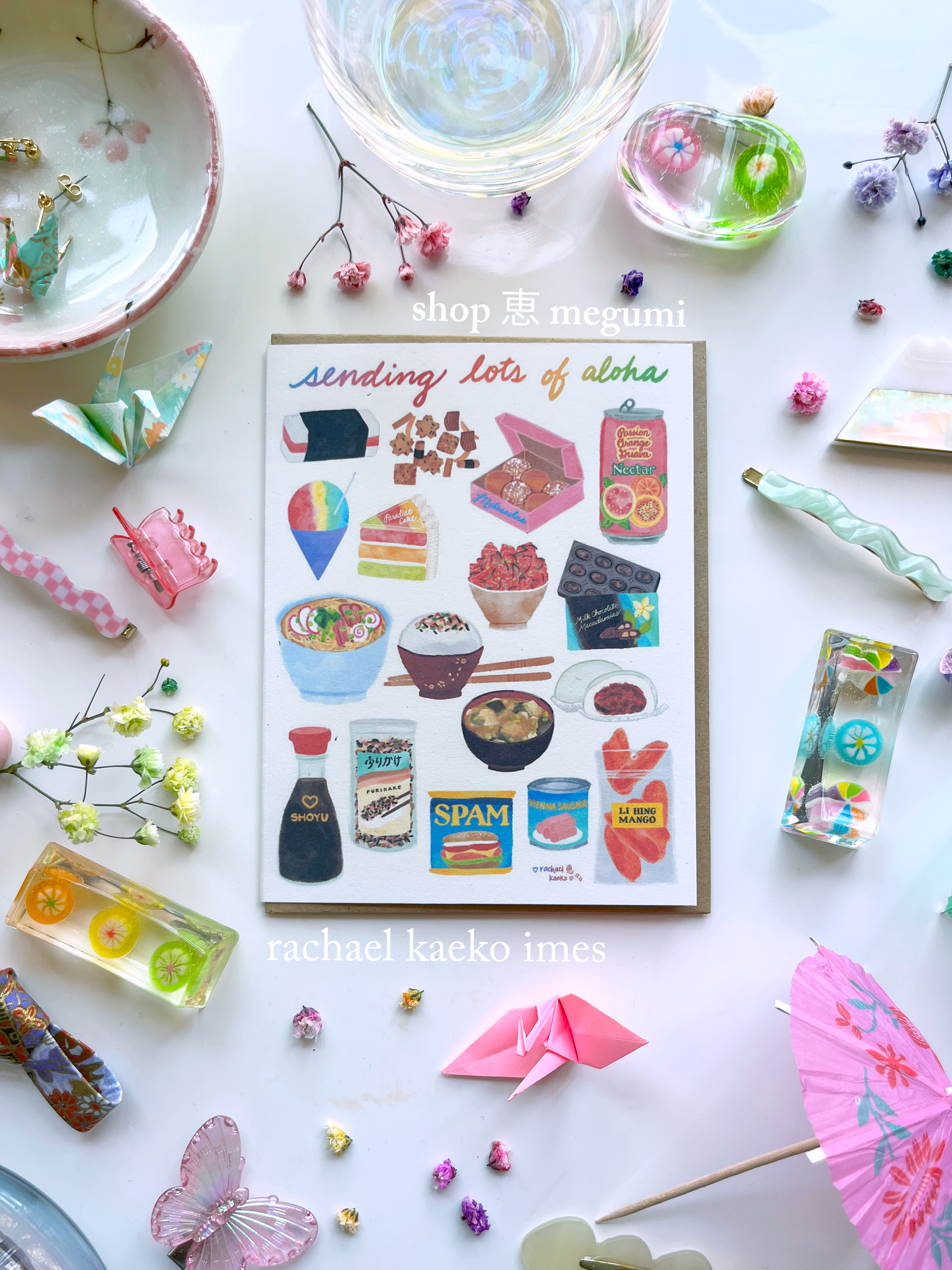 Hawaii-inspired greeting card that reads "Sending Lots of Aloha" in cursive text, with colorful illustrations of Hawaii-style foods, on a white surface with colorful decorative objects.