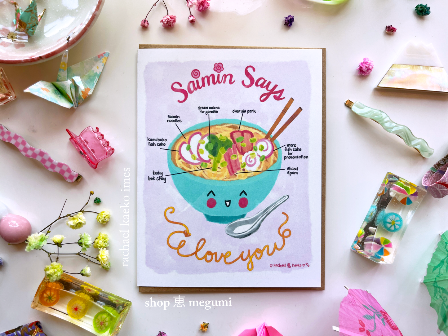 Hawaii-inspired greeting card with 'Saimin Says I Love You' illustrated text design and illustration of a smiling Hawaiian saimin ramen bowl on white surface with decorative objects.