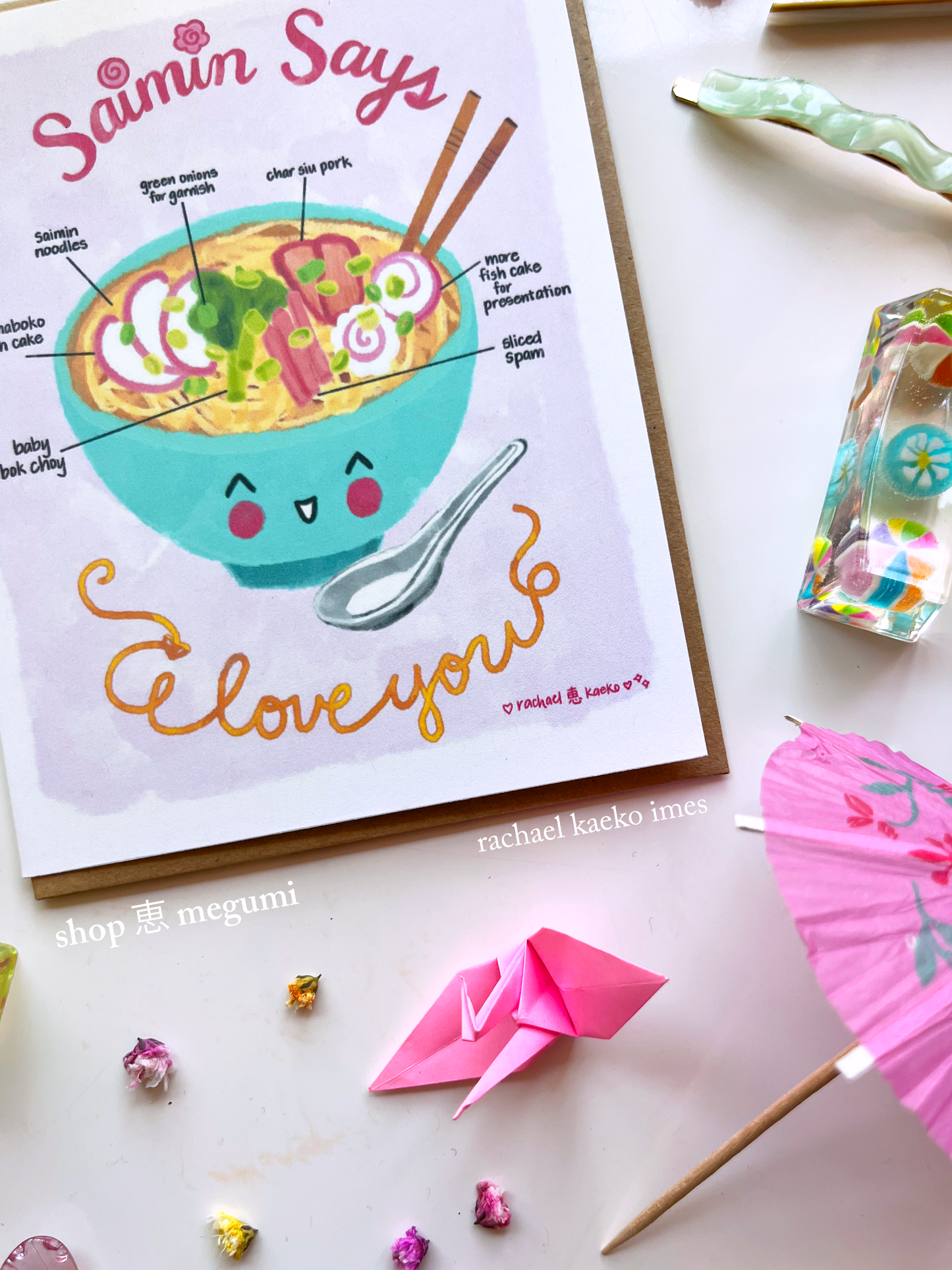 Close-up of Hawaii-inspired greeting card, with a cute illustrated bowl of Hawaiian saimin ramen and text on a white surface with decorative items.
