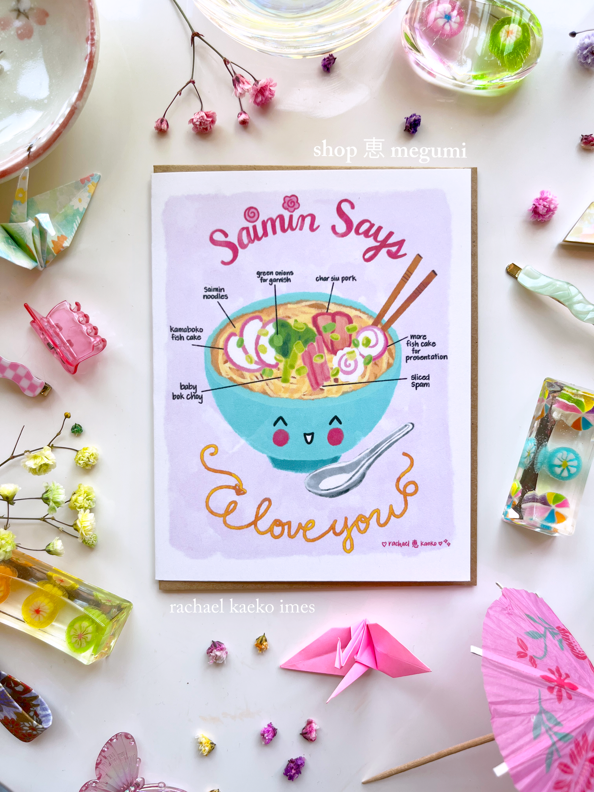 Hawaii-inspired greeting card with 'Saimin Says I Love You' illustrated text design and illustration of a smiling Hawaiian saimin ramen bowl on white surface with decorative objects.