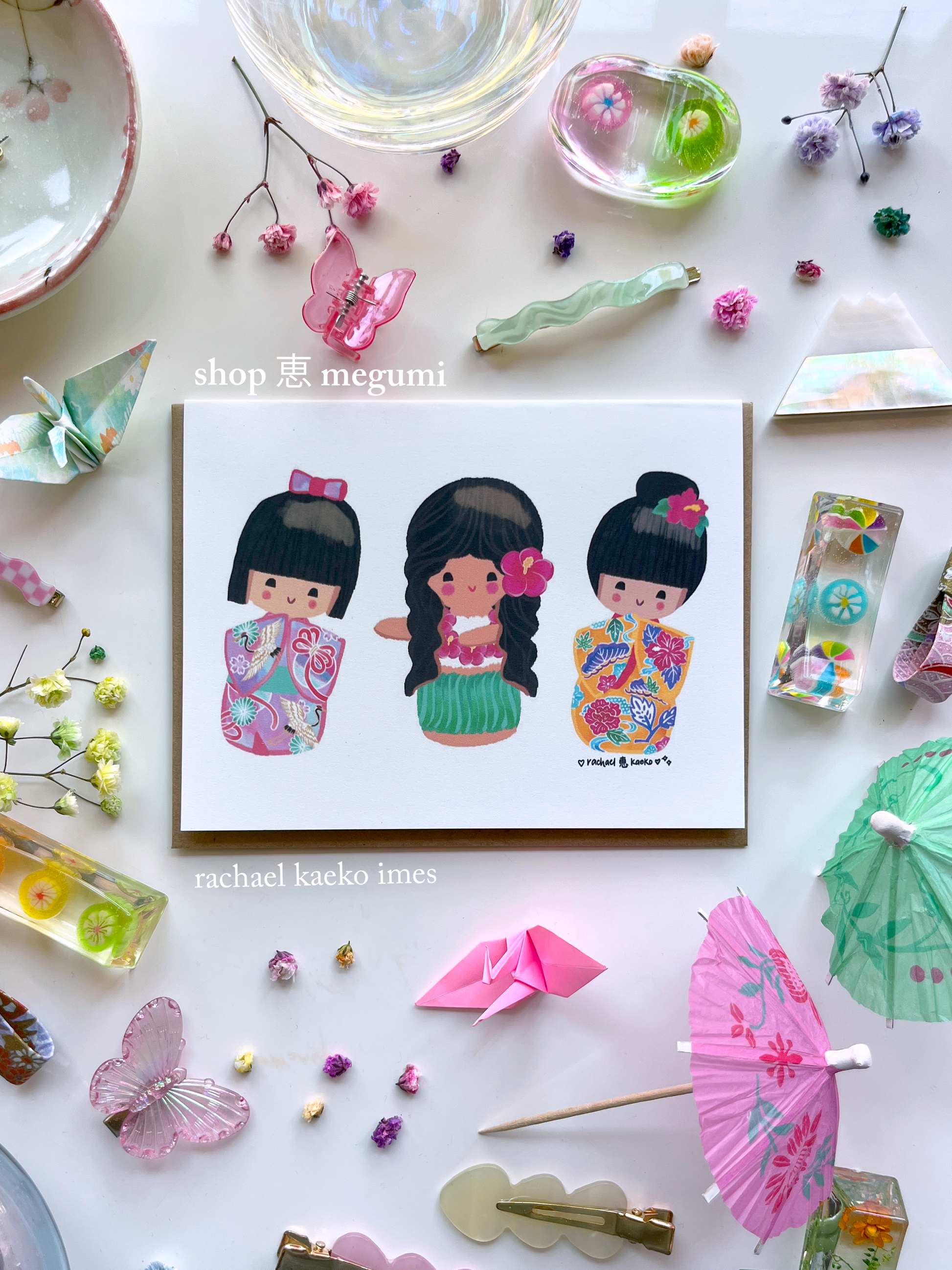 Colorful greeting card of original shop megumi kokeshi doll girls in traditional cultural outfits on a white surface with decorative items.