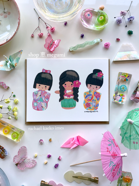 Colorful greeting card of original shop megumi kokeshi doll girls in traditional cultural outfits on a white surface with decorative items.