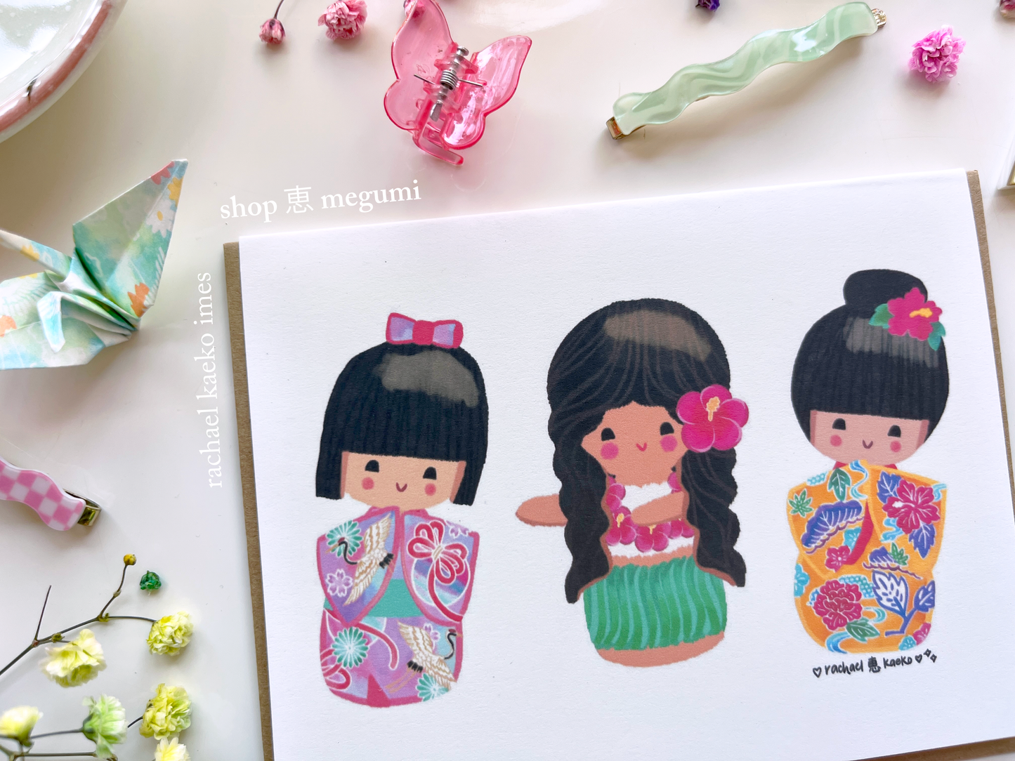 Close up image of a colorful greeting card with original shop megumi kokeshi doll character illustrations on a white background with decorative objects.