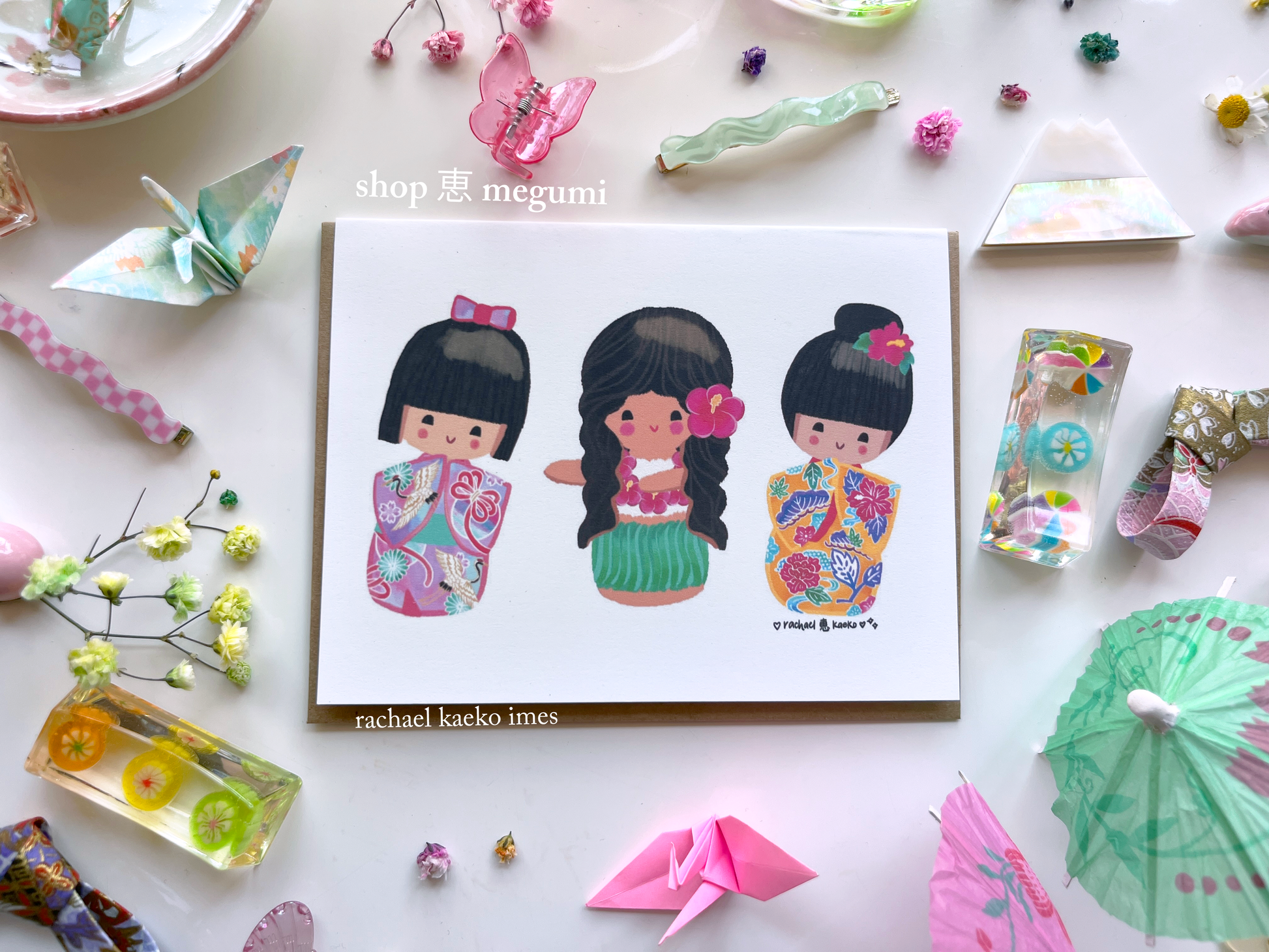 Colorful greeting card of original shop megumi kokeshi doll girls on a white surface with origami and flowers.