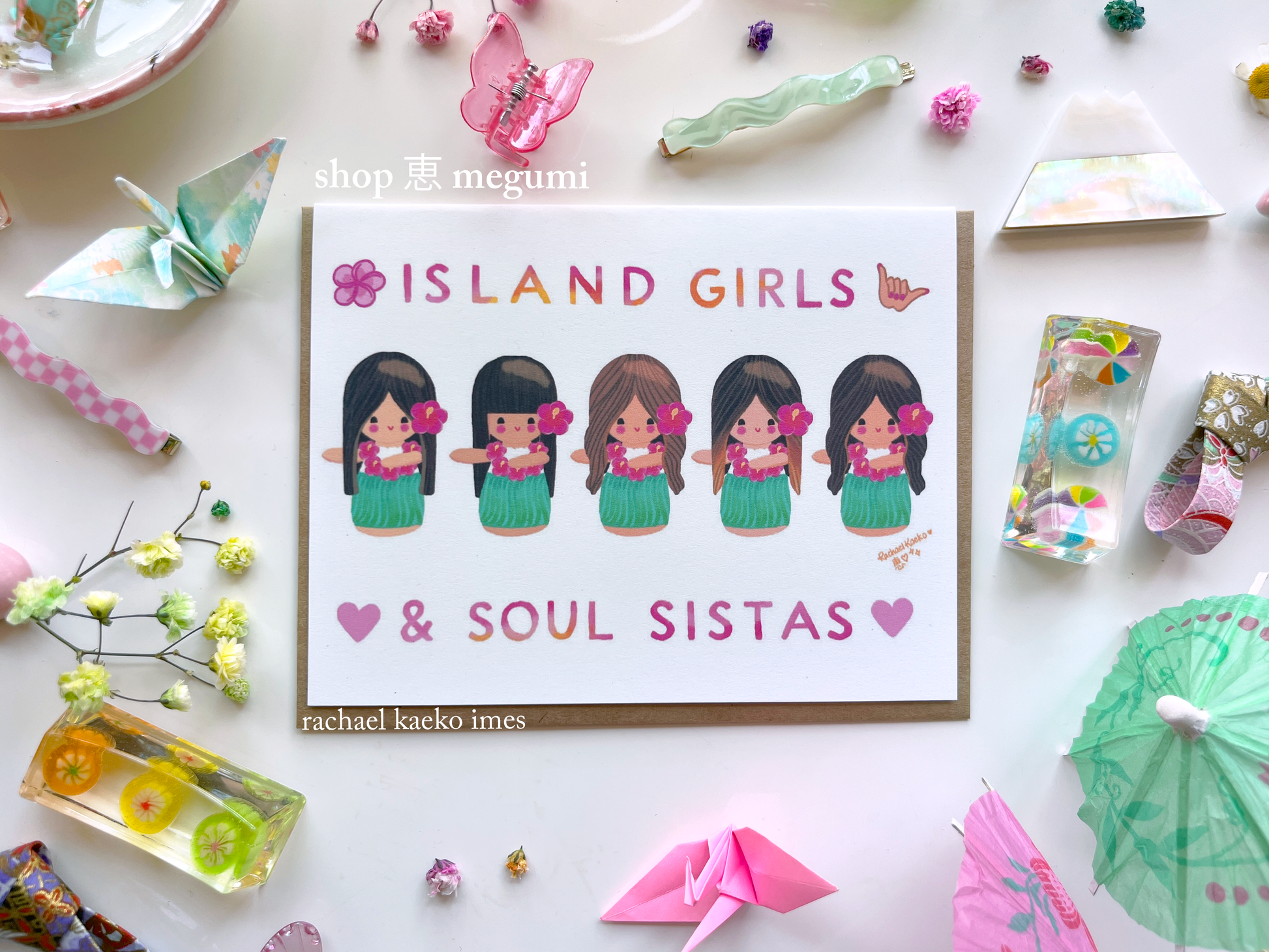 Colorful Hawaii-inspired greeting card featuring original Shop Megumi kokeshi hula girls illustrations, with text that reads "Island Girls & Soul Sistas", on a white surface with decorative items.