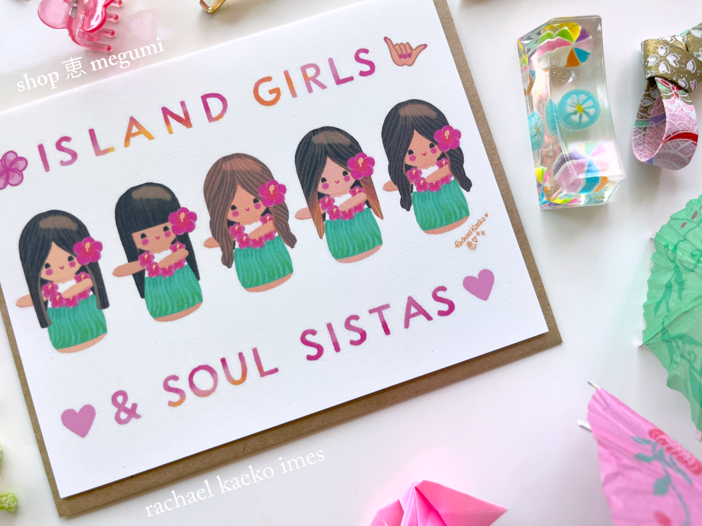 Colorful Hawaii-inspired greeting card featuring original Shop Megumi kokeshi hula girls illustrations, with text that reads "Island Girls & Soul Sistas", on a white surface with decorative items.