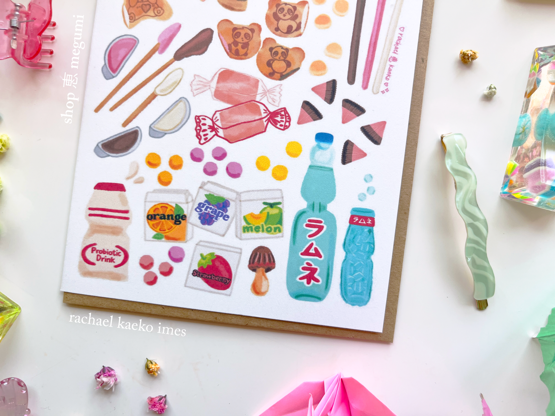 Close-up of Japanese-inspired greeting card with colorful illustrations of Japanese snacks, on a white surface, surrounded by decorative items.