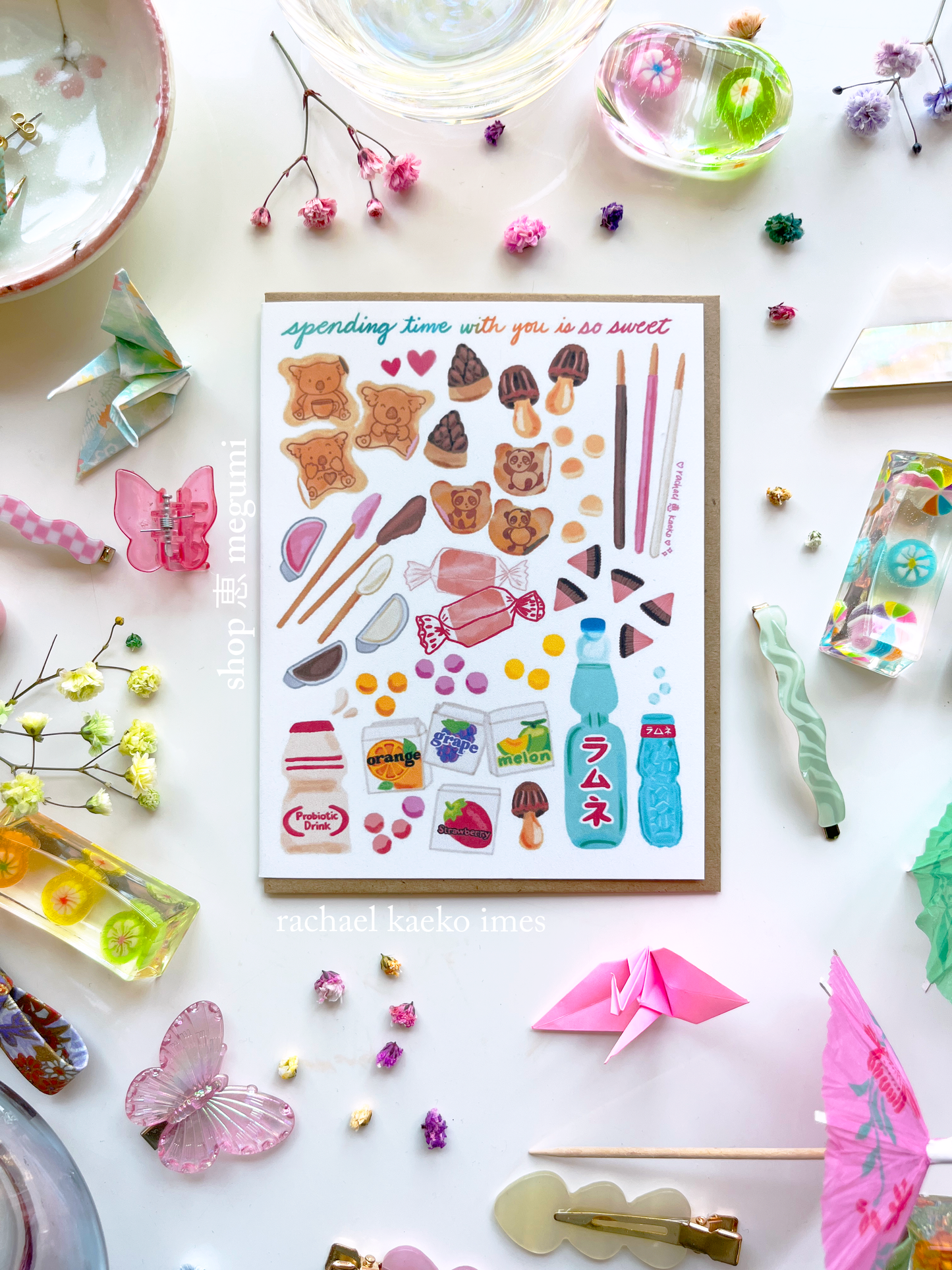 Japanese-inspired greeting card with text that reads "Spending Time with You is so Sweet" and colorful illustrations of Japanese snacks and treats, on a white surface surrounded by decorative objects.