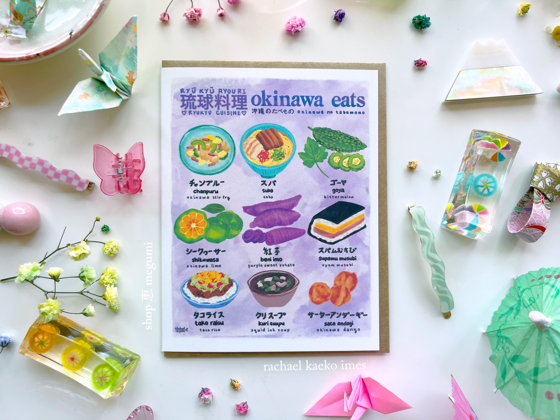 "Okinawa Eats" greeting card with colorful illustrations of Okinawan cuisine on a white surface with decorative items.