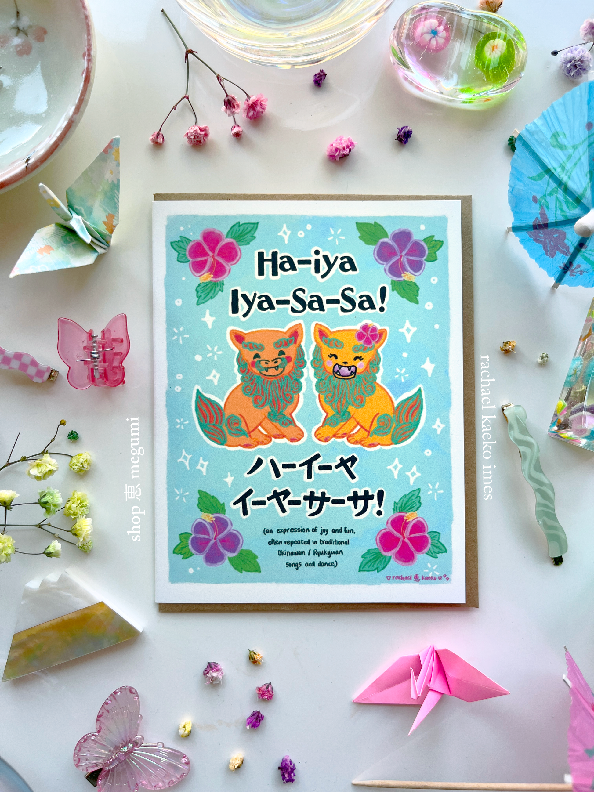 Okinawa-inspired greeting card with 'Ha-ya Iya-Sa-Sa!' text and colorful illustrations of Okinawan shiisa lions and hibiscus flowers, on a decorative surface with origami and flowers.