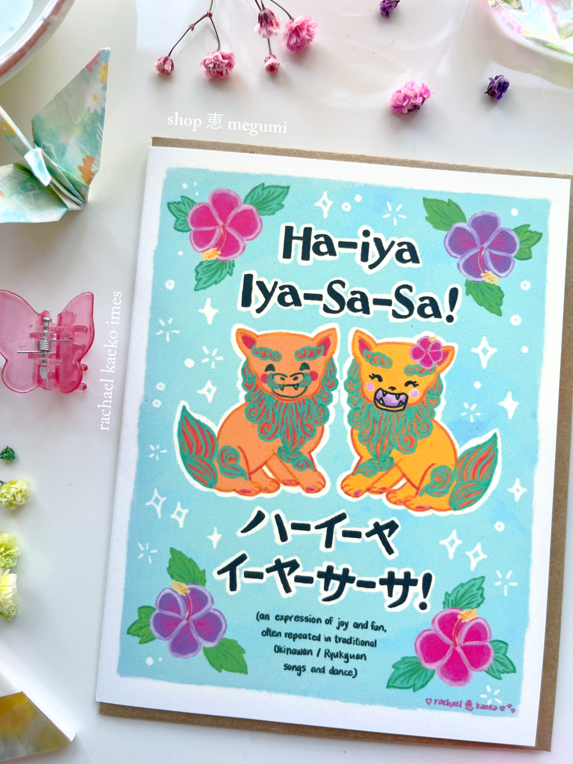 Greeting card with 'Ha-iya Iya-Sa-Sa!' text and colorful illustrations of Okinawa shiisa lions and hibiscus flowers, on a white surface.