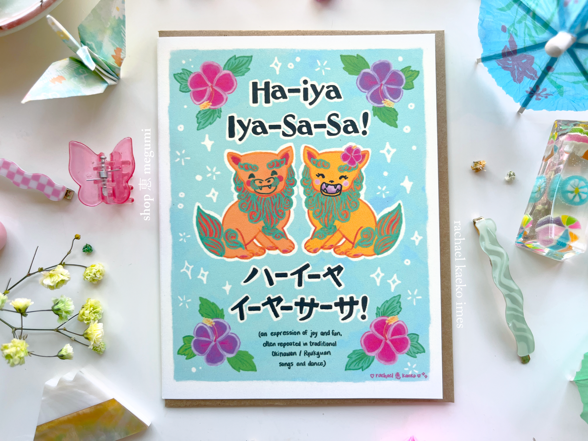 Okinawa-inspired greeting card with two orange Okinawa shiia lions, and Japanese and English text that reads, 'Ha'iya 'iya sa sa!' over a blue background, on a white surface surrounded by decorative items.