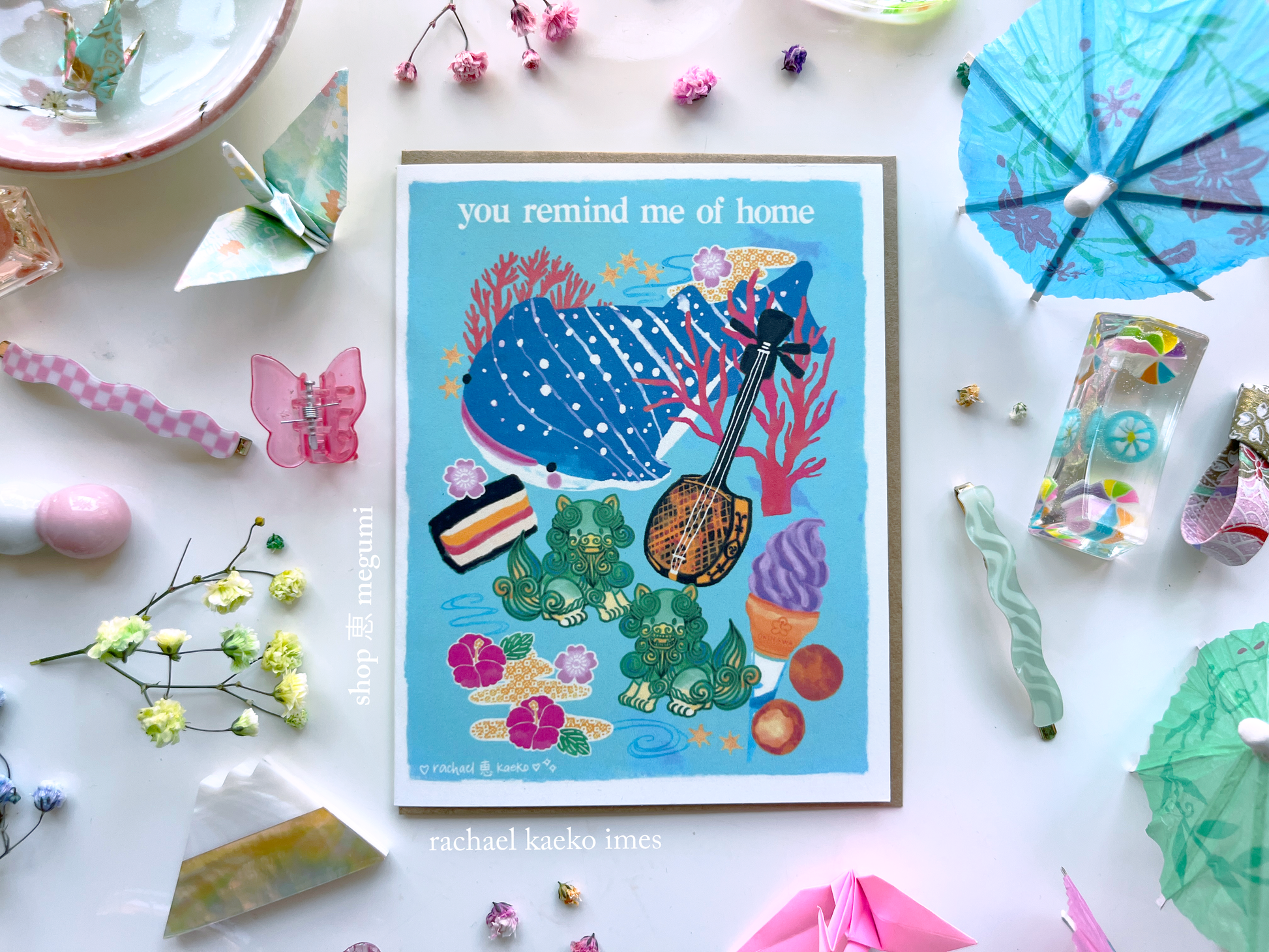 Okinawa-inspired greeting card with illustrated images of Okinawan culture, including a blue spotted whale shark, colorful coral and star-shaped sand, hibiscus flowers and bingata patterns, shiisa lions, beni imo ice cream, Okinawan andagi dango, and spam musubi, on a white surface, surrounded by colorful decorative items.