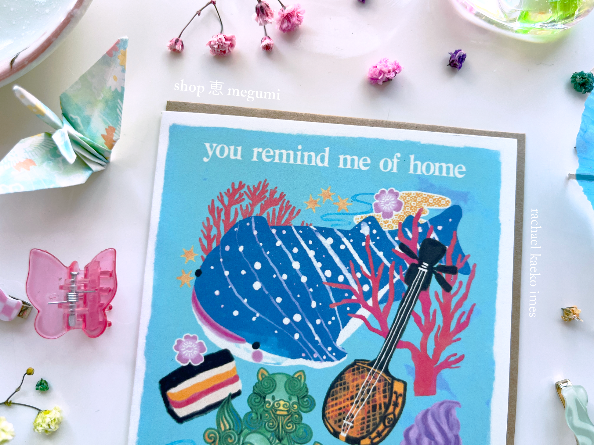 Close up image of Okinawa-inspired greeting card, with blue spotted whale shark and coral illustrations, with text that reads "You Remind Me of Home", on a white surface surrounded by colorful decorative objects.