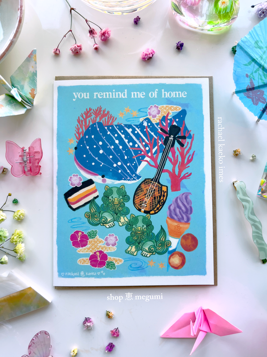 Okinawa-inspired greeting card with illustrated images of Okinawan culture, including a blue spotted whale shark, colorful coral and star-shaped sand, hibiscus flowers and bingata patterns, shiisa lions, beni imo ice cream, Okinawan andagi dango, and spam musubi, on a white surface, surrounded by colorful decorative items.