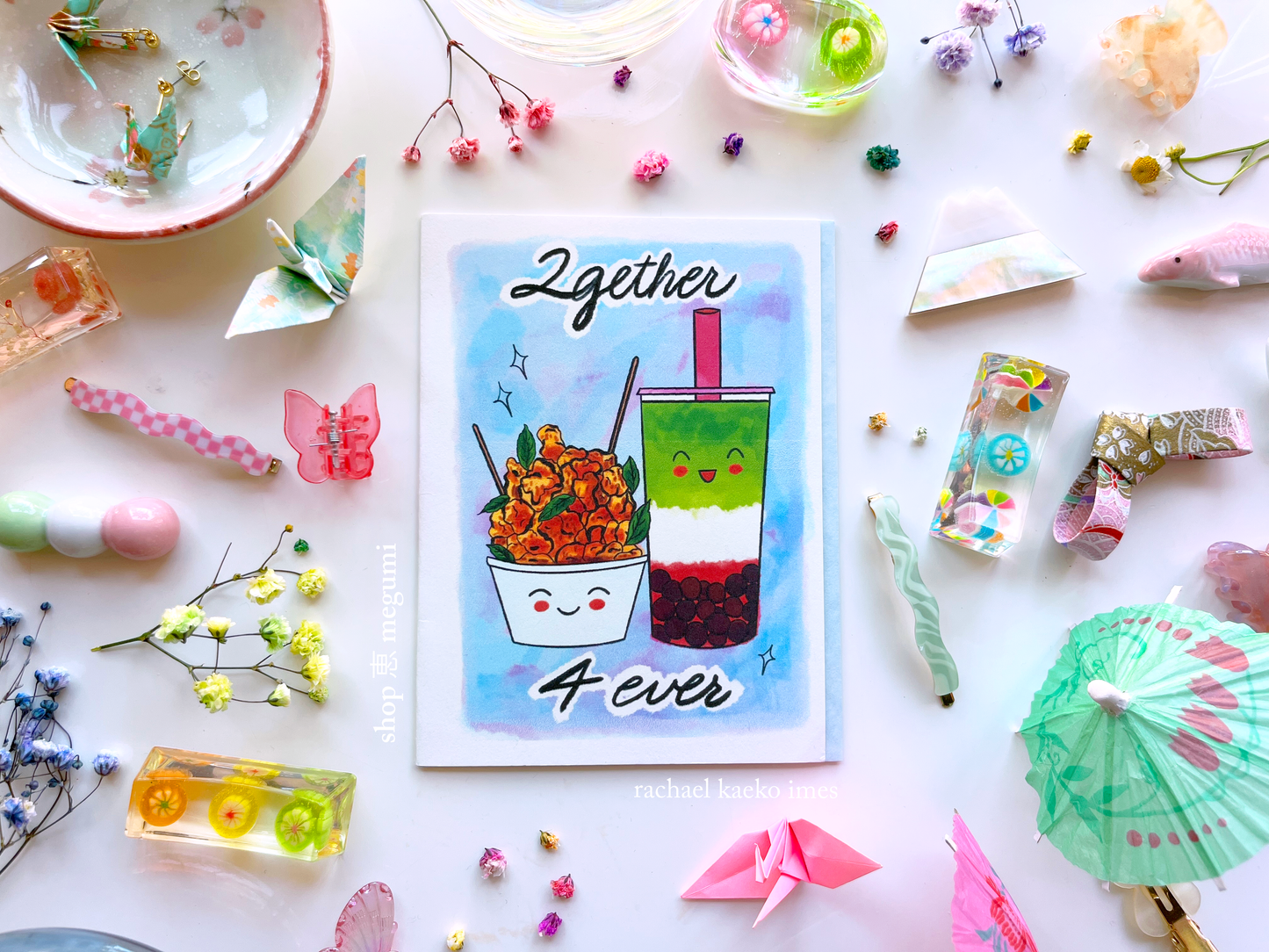 Boba-inspired greeting card with text that reads '2gether 4ever', and illustration of a smiling strawberry matcha boba next to a smiling bowl of popcorn chicken, on a white surface surrounded by decorative items.