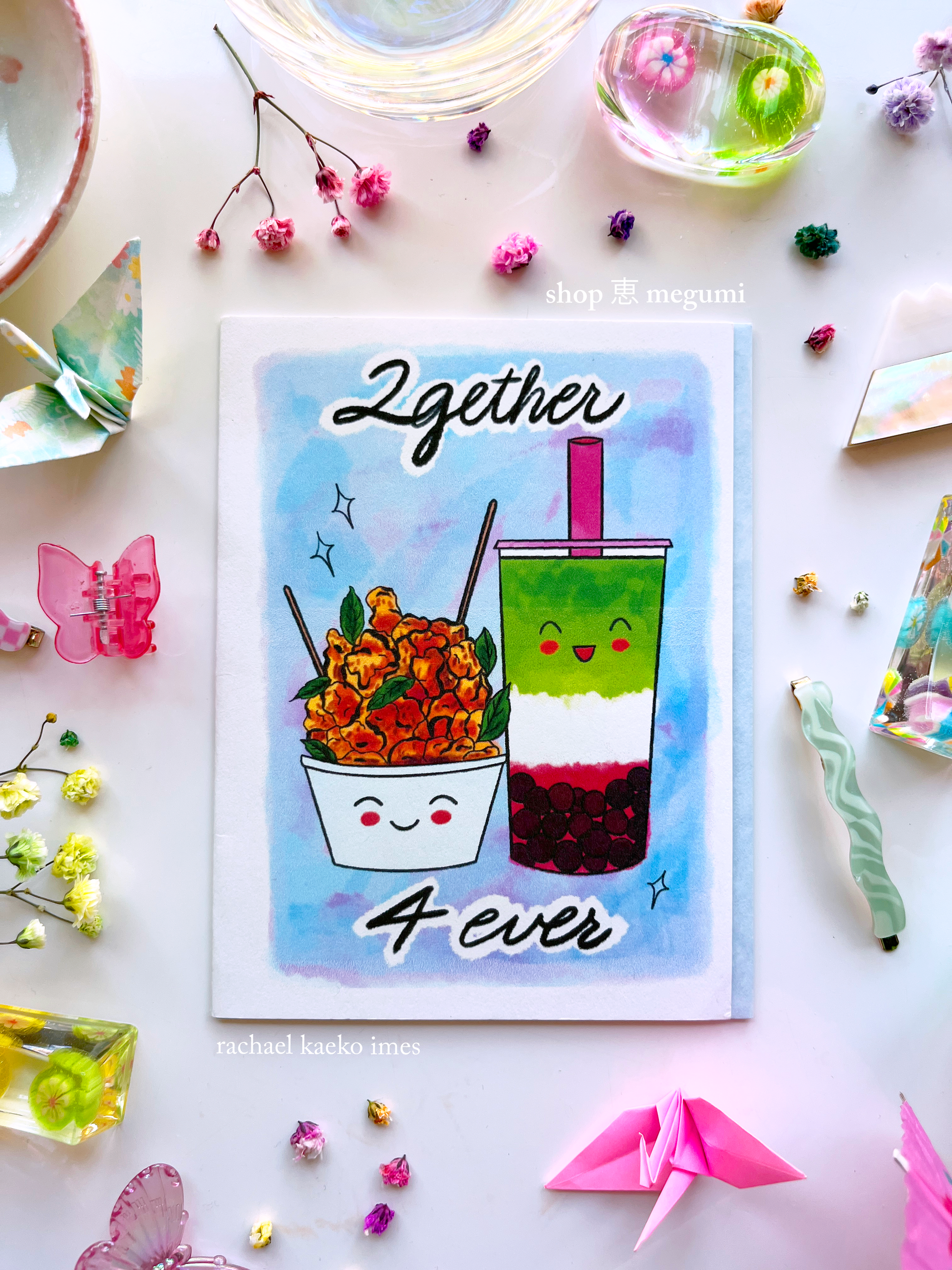 Boba-inspired greeting card with text that reads '2gether 4ever', and illustration of a smiling strawberry matcha boba next to a smiling bowl of popcorn chicken, on a white surface surrounded by decorative items.