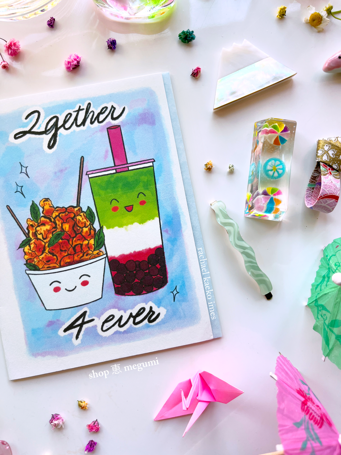 Close-up image of boba-inspired greeting card with a design of a smiling strawberry matcha boba and popcorn chicken, with illustrated '2gether 4 ever' text, surrounded by decorative items on a light background.